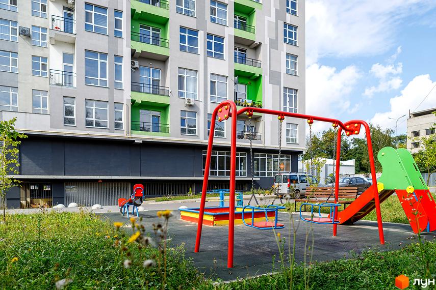 Житловий комплекс ЕкоПарк: сіра багатоповерхівка з зеленими елементами та дитячим майданчиком зі гойдалками та гіркою.