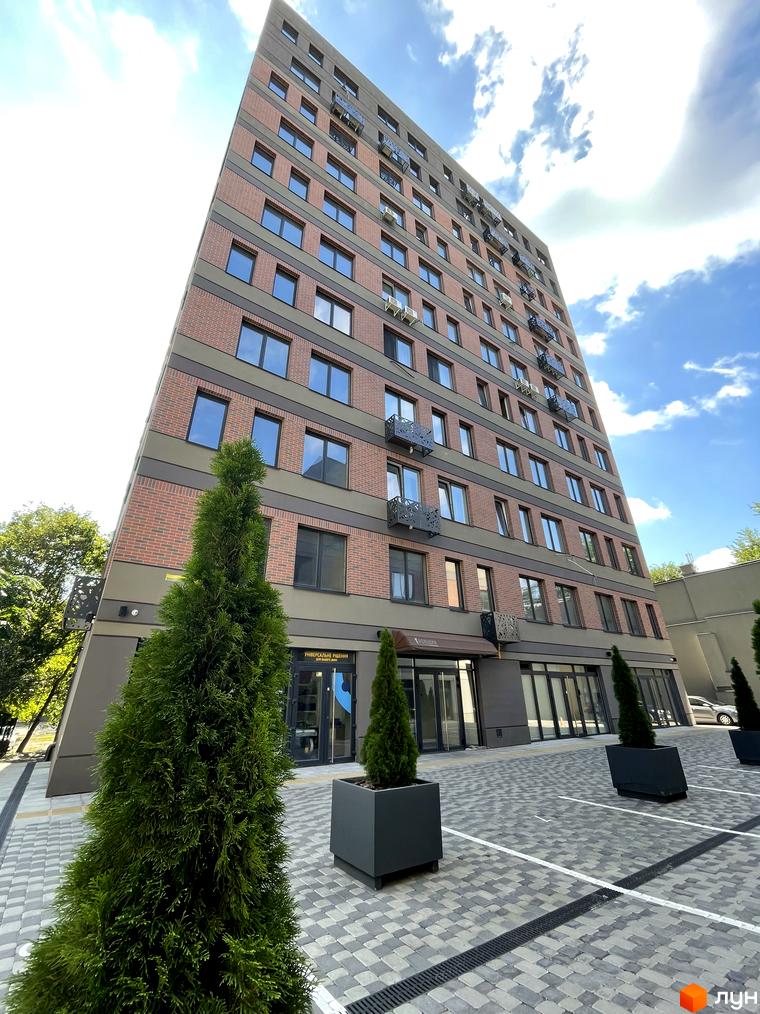 Висотна будівля ЖК Троїцький з цегляним фасадом та панорамними вікнами. На передньому плані доглянута прибудинкова територія з декоративними деревами.