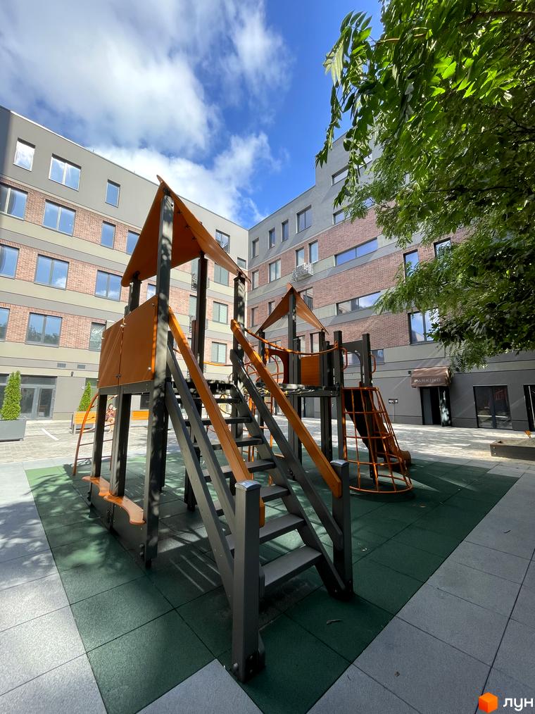 Житловий комплекс Троїцький. Фасад будинку з цегли та сірого кольору. Дитячий майданчик з гіркою та сходами на території.