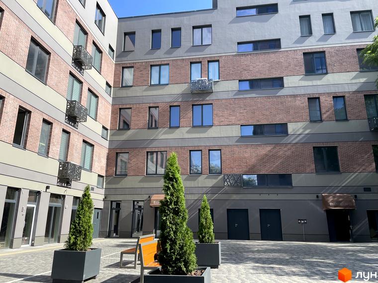 Зображення сторінки житлового комплексу Троїцький. Багатоповерховий будинок з цегляною кладкою та великими вікнами. На території комплексу є зелені насадження та лавки для відпочинку.