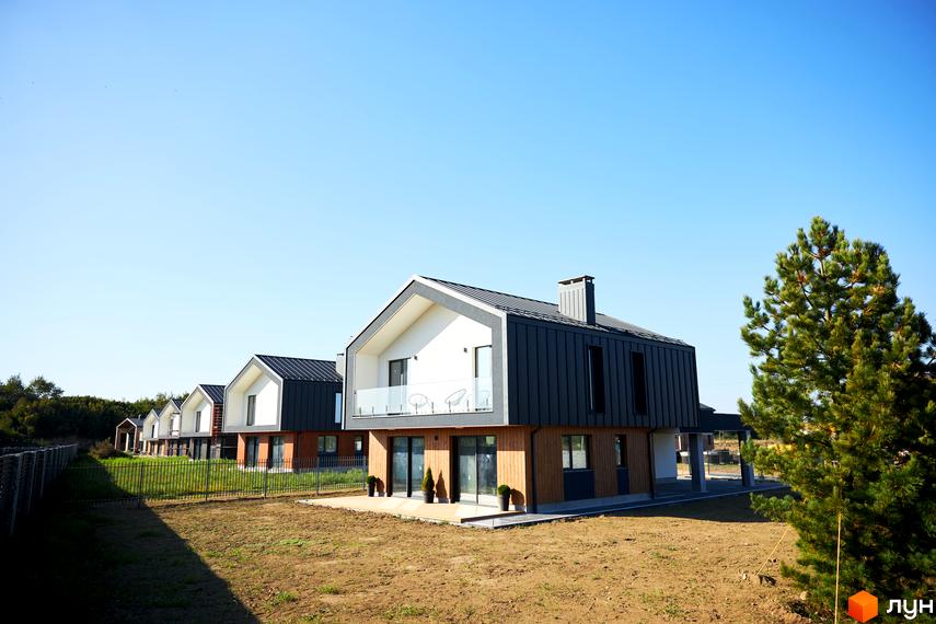 Котеджне містечко OZON Village: двоповерхові будинки з темними дахами та дерев'яними фасадами на тлі блакитного неба. Навколо будинків зелена трава та дерева.
