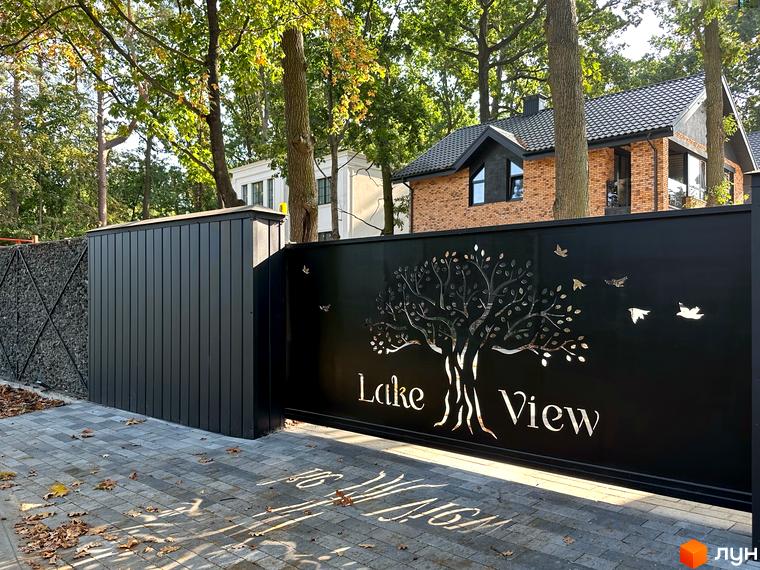 Котеджне містечко Lake View. Чорний кований паркан з витіюватим деревом та птахами, вимощені сірою плиткою доріжки та зелені дерева навколо.