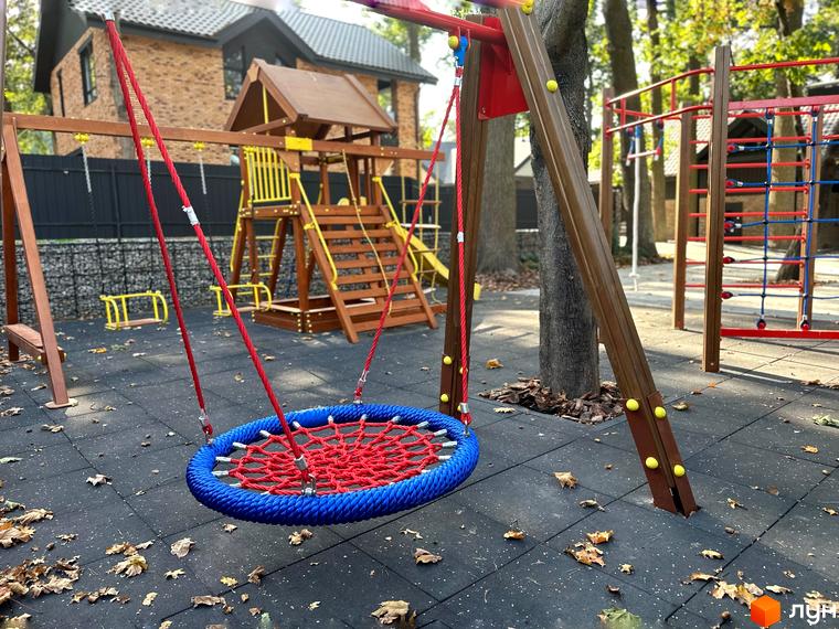 Дитячий майданчик біля будинку КМ Lake View з гойдалками та гірками. Територія комплексу упорядкована, з безпечним покриттям та зеленими насадженнями.