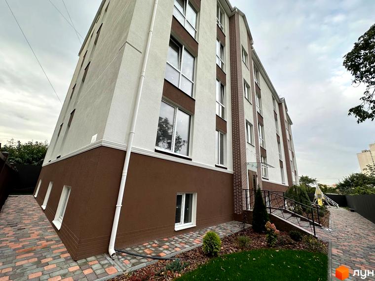 Зовнішній вигляд новобудови ЖК SMART IDEA з коричневим цоколем та білими стінами. Прибудинкова територія з бруківкою та зеленими насадженнями.