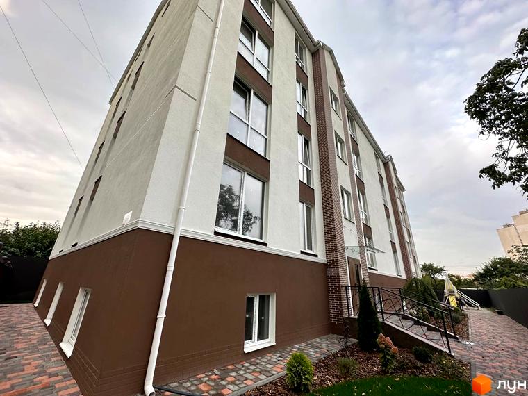 Зовнішній вигляд новобудови ЖК SMART IDEA з коричневим та білим фасадом. Навколо будинку розташована бруківка та зелені насадження.