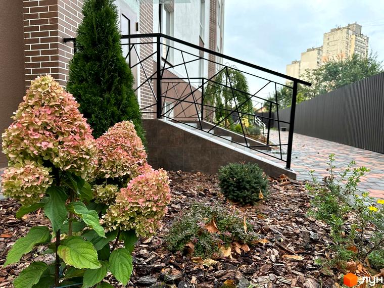 Зовнішній вигляд житлового комплексу SMART IDEA з доглянутою прибудинковою територією та квітучими рослинами. На фото видно цегляний фасад та сучасну огорожу.