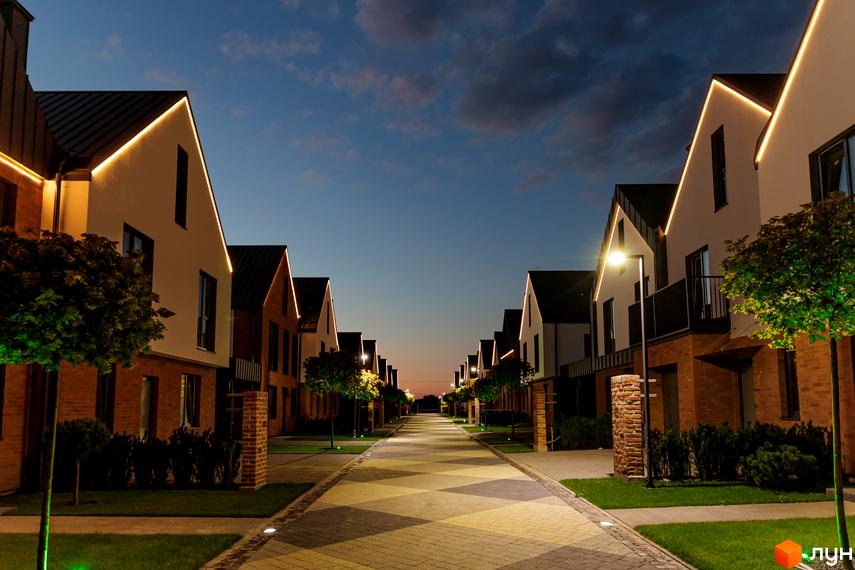 Котеджне містечко КМ LAGOM: фасади будинків з підсвічуванням та доглянута прибудинкова територія з деревами та бруківкою.