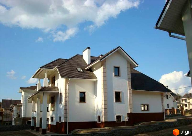 Фото Котеджного містечка Іванковичі. Двоповерховий будинок з білими стінами та коричневим дахом на фоні блакитного неба.