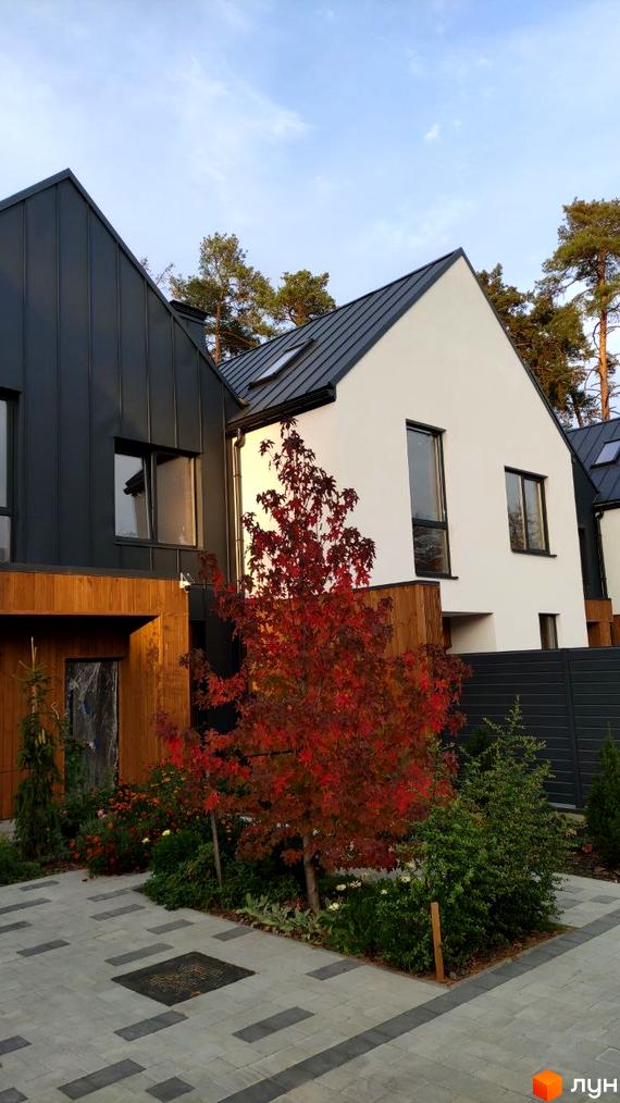 Котеджне містечко КМ Toscana House 2 з білою та сірою фасадною обробкою, металевим дахом та доглянутою прибудинковою територією з деревами та квітами.