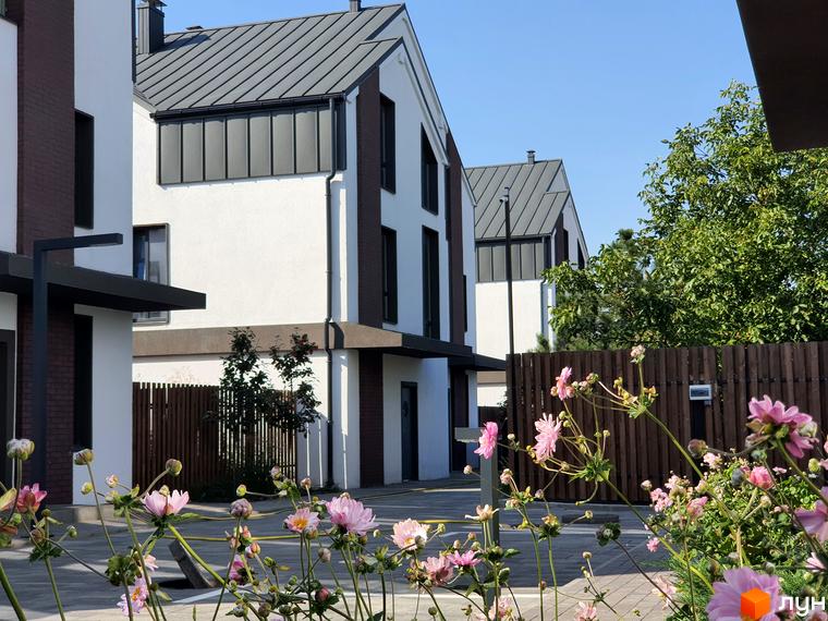 Зовнішній вигляд таунхаусів New Smart 6 з білою та коричневою обробкою фасаду та сірим дахом. На прибудинковій території видно зелені дерева та квіти.