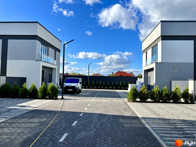 Зовнішній вигляд будинків КМ GREAT OSOCOR Apartments з сірими та білими фасадами, скляними балконами та акуратною прибудинковою територією з туями та бруківкою.