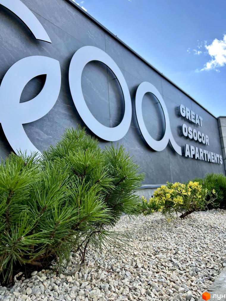Фасад комплексу GREAT OSOCOR Apartments з великими білими літерами на сірій стіні. Попереду зелені ялинки та біла галька.