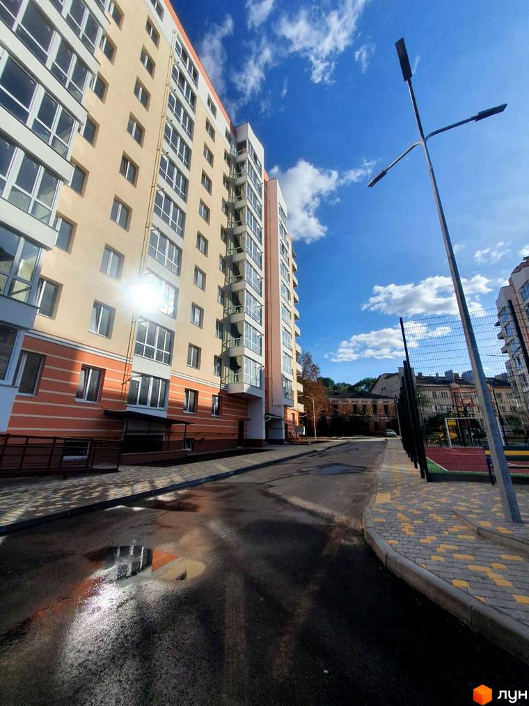 ЖК Зоря з бежевими фасадами, балконами та сонячними відблисками. На прибудинковій території видно дорогу, тротуар, спортивний майданчик та калюжі.