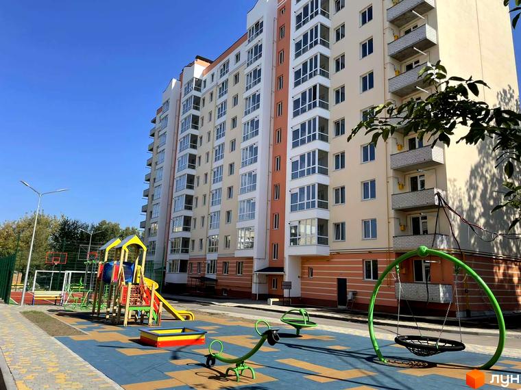 Новобудова Зоря: багатоповерховий будинок з бежевими стінами та помаранчевими акцентами. Поруч облаштований дитячий майданчик з гойдалками та гіркою.