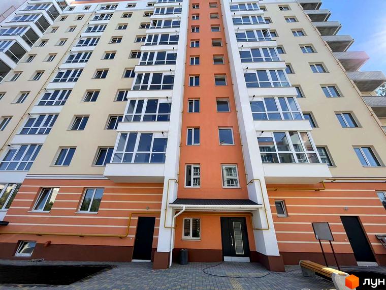 Багатоповерховий будинок Житловий комплекс Зоря з бежевим та помаранчевим фасадом, великими вікнами. Перед будинком вимощена територія з лавкою.