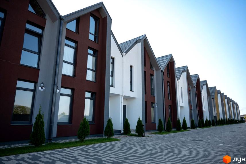 Котеджне містечко КМ Beverly Hills Residence: ряд сучасних будинків з коричневою та білою обробкою, сірими дахами та великими вікнами. Прибудинкова територія з акуратними ялинками та бруківкою.