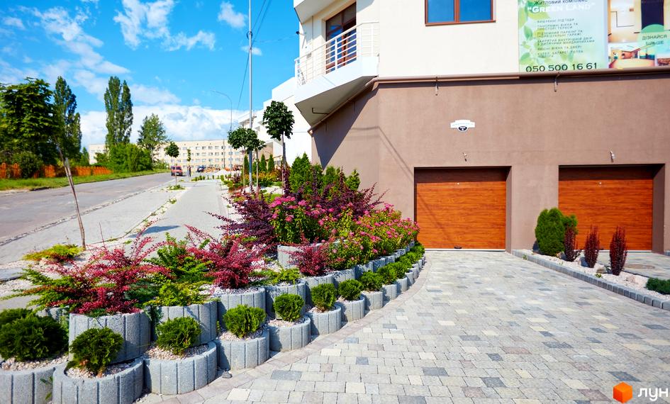 Житловий комплекс GREEN LAND з двома дерев'яними гаражними воротами та доглянутою прибудинковою територією з квітучими рослинами та кущами.