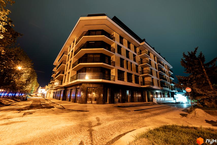 Новобудова ЖК RIVER LAND з балконами та панорамними вікнами вночі. Навколо будинку освітлена вулиця з деревами та бруківкою.