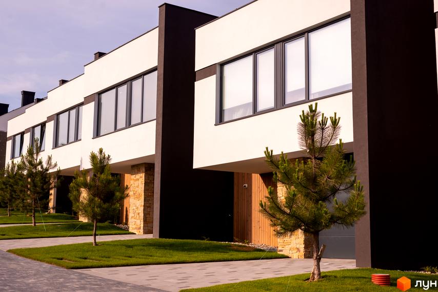 Котеджне містечко КМ SHERWOOD з біло-темними фасадами та великими вікнами. Озеленена територія з молодими соснами та доріжками з плитки.