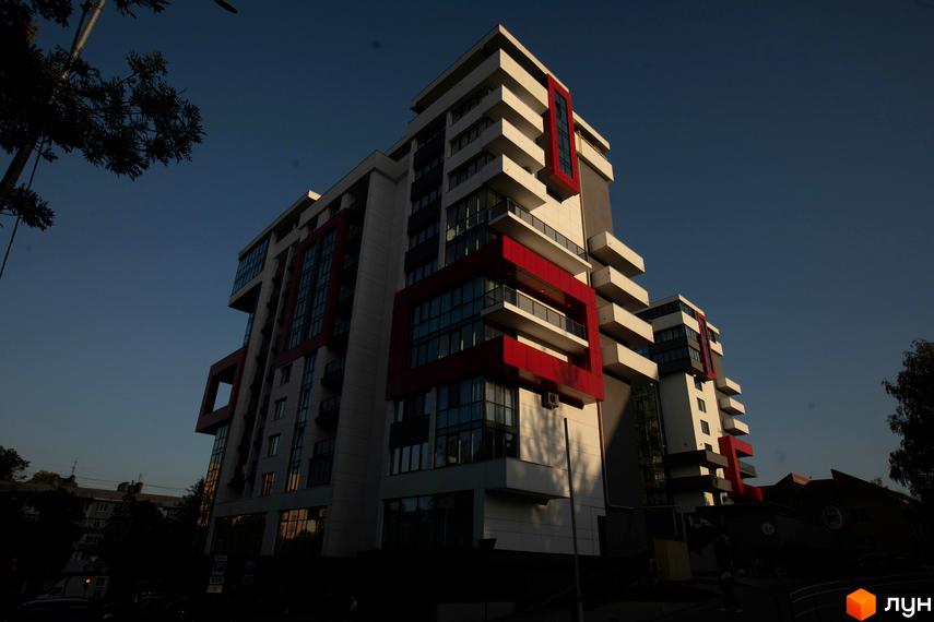 Житловий комплекс Cube House з білим фасадом, червоними акцентами та великими вікнами. Будівля розташована на фоні блакитного неба, поруч видно дерева та дорогу.