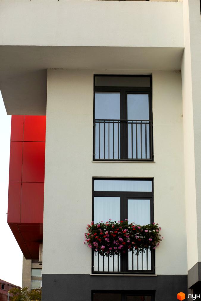 Фасад житлового комплексу Cube House з білими стінами, червоними архітектурними елементами та балконами. На нижньому балконі видно квітковий ящик з різнокольоровими квітами.