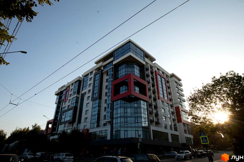 Багатоповерховий будинок Житлового комплексу Cube House з великими вікнами та червоними акцентами. Навколо будинку проїжджа частина з автомобілями та зелені дерева.