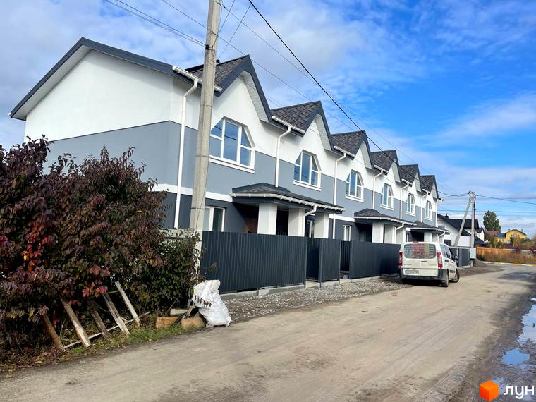 Таунхауси „Комфортне містечко“: ряд двоповерхових будинків біло-сірого кольору з темними дахами та арочними вікнами. Перед ними ґрунтова дорога та темні огорожі.