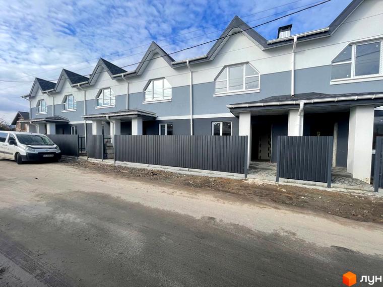 Таунхауси „Комфортне містечко“: ряд сіро-білих будинків з гостроверхими дахами. Перед ними темні огорожі та ґрунтова дорога.