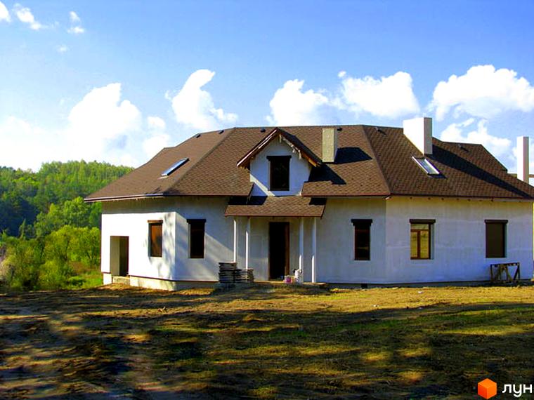 Зображення будинку в Котеджному містечку Лісове озеро. Двоповерховий будинок з коричневою черепицею та білими стінами на тлі зеленого лісу.