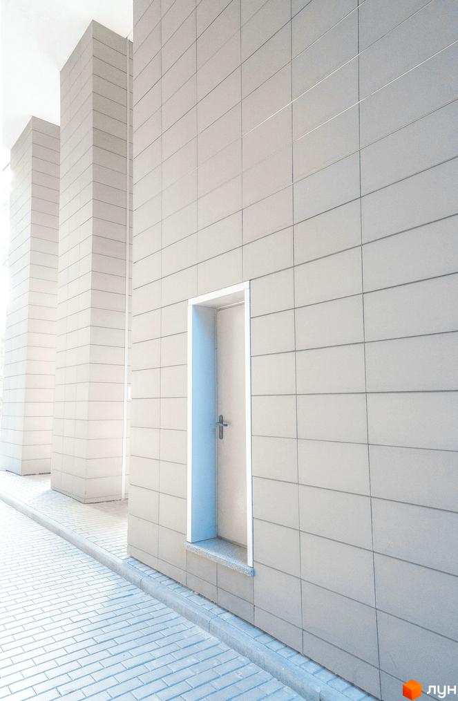 Зовнішній вигляд новобудови ЖК Crystal Park Tower з білими колонами та сірою плиткою. Поруч бруківка та сірий бордюр.