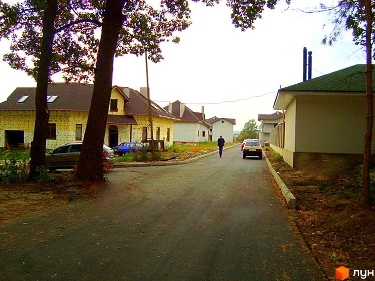 Зображення Котеджного містечка Лісове озеро: одноповерхові будинки з коричневими дахами вздовж асфальтованої дороги. Навколо будинків дерева та припарковані автомобілі.