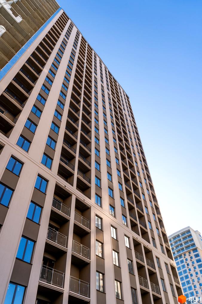 Висотна будівля ЖК Еллада з великою кількістю вікон та балконів на фоні блакитного неба. Архітектура житлового комплексу з прямими лініями та чіткими формами.