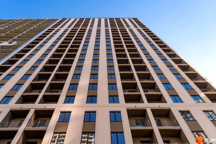 Висотна новобудова ЖК Еллада з великою кількістю балконів та вікон на тлі блакитного неба. Фасад будівлі виконаний у світлих тонах.