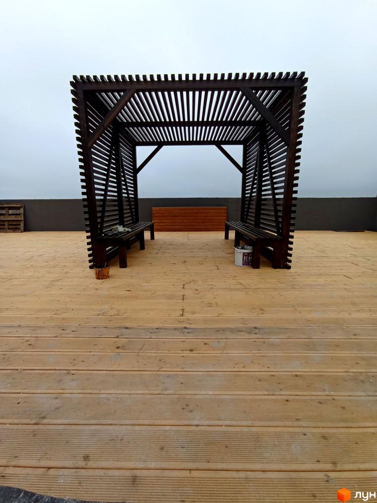 Відкрита дерев'яна альтанка з лавками на дерев'яному настилі. Територія навколо альтанки вкрита дерев'яними дошками.