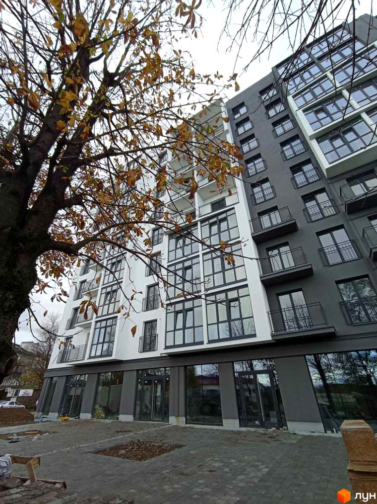 Зовнішній вигляд житлового комплексу Сонячна Долина з великою кількістю вікон та балконів, поруч дерево з осіннім листям. Прибудинкова територія з бруківкою та будівельними матеріалами.