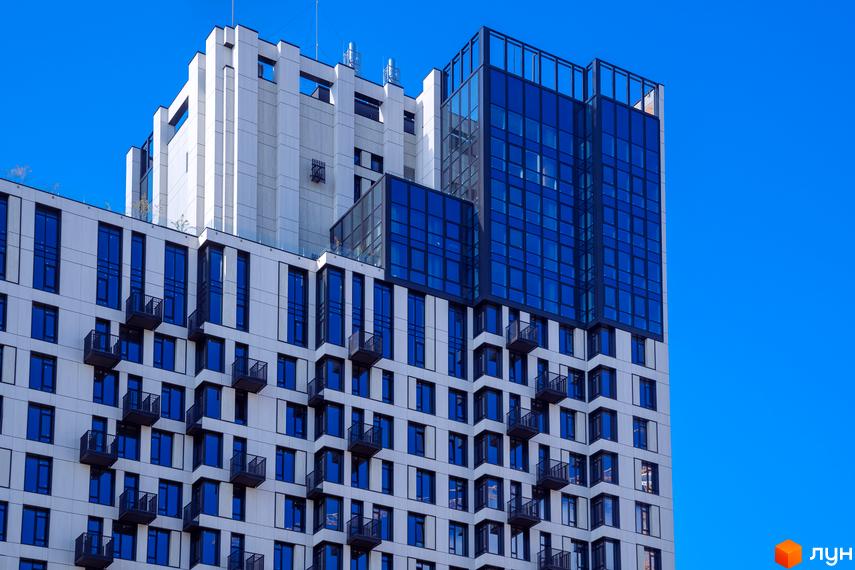 Сучасний житловий комплекс TRIIINITY з біло-сірим фасадом та синіми вікнами. Будівля має багато балконів та скляні елементи на верхніх поверхах.