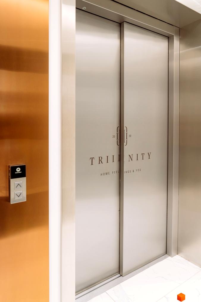 Інтер'єр ліфта в житловому комплексі TRIIINITY з дверима з нержавіючої сталі та логотипом проєкту. Поруч розташована панель керування ліфтом.