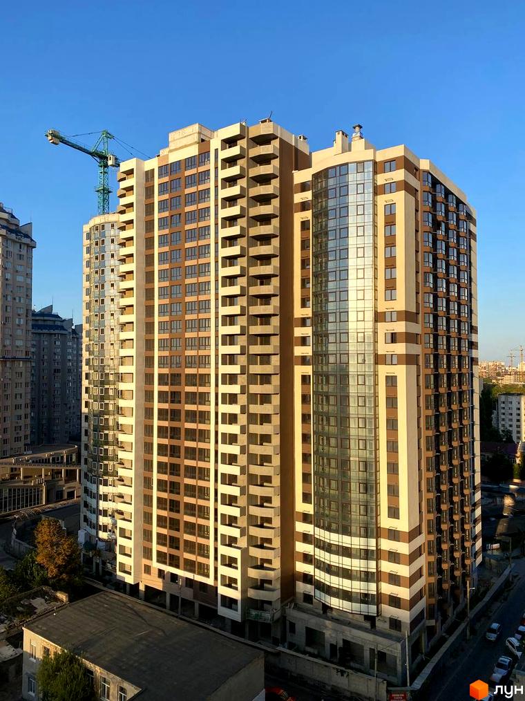 Висотна новобудова з великою кількістю вікон та балконів. На території комплексу є зелені насадження та парковка для автомобілів.