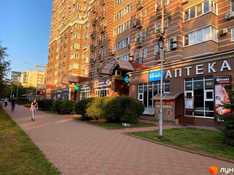 Житловий комплекс Голосіїво: багатоповерховий будинок з цегляним фасадом та комерційними приміщеннями на першому поверсі, поруч брукована доріжка та зелені насадження.