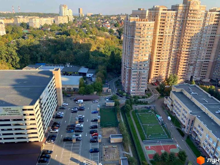 Високі житлові будинки Комплексу Голосіїво з багатьма вікнами. Поруч розташовані багаторівневий паркінг, відкрита автостоянка та спортивні майданчики.