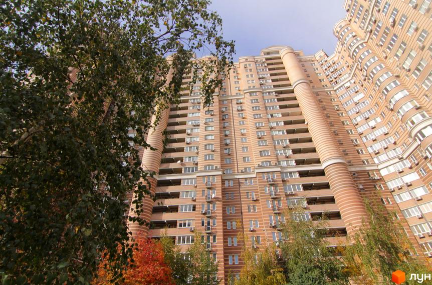 Багатоповерховий житловий комплекс Голосіїво світло-коричневого кольору з численними балконами. На передньому плані видно дерева із зеленим та осіннім листям.