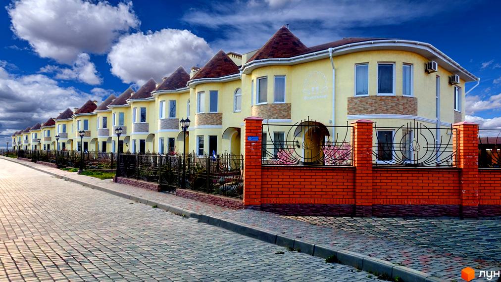 Зовнішній вигляд житлового комплексу КМ Перлинне з жовтими будинками та червоними цегляними огорожами. Прибудинкова територія з тротуарною плиткою та ліхтарями.