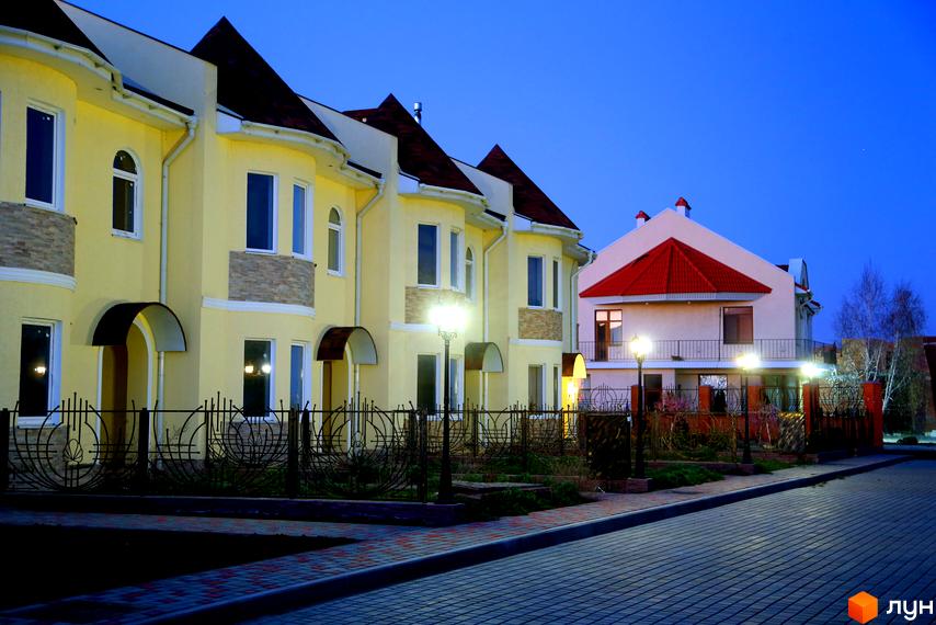 Зовнішній вигляд житлового комплексу КМ Перлинне з жовтими будинками та червоним дахом. Прибудинкова територія з освітленням та бруківкою.