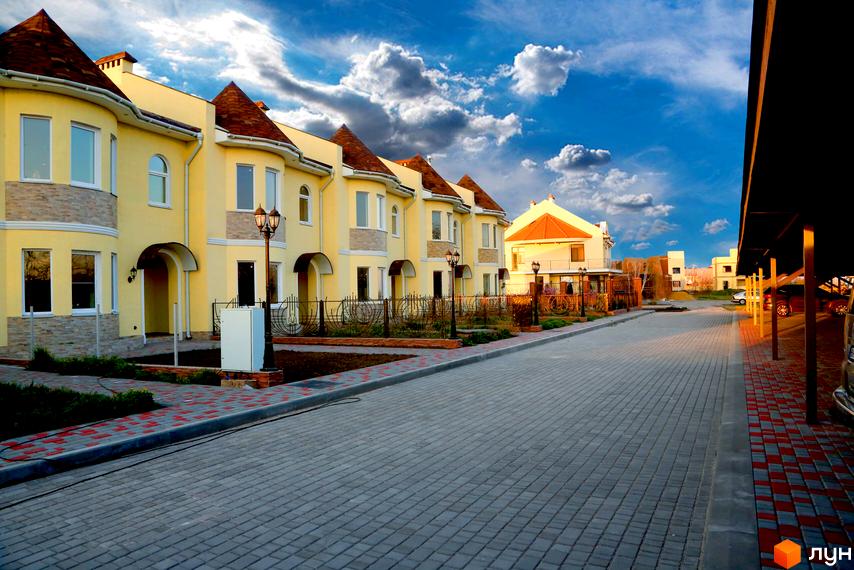 Зображення Котеджного містечка Перлинне: жовті двоповерхові будинки з коричневими дахами вздовж вулиці з тротуарною плиткою. Навколо будинків зелені насадження та декоративні огорожі.