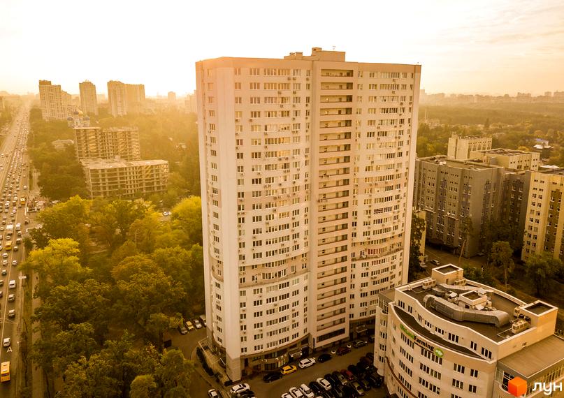 Житловий комплекс Вікторія – висотна будівля світлого кольору з численними вікнами та балконами. Поруч розташовані зелені насадження, міська дорога з рухом та інші будинки.