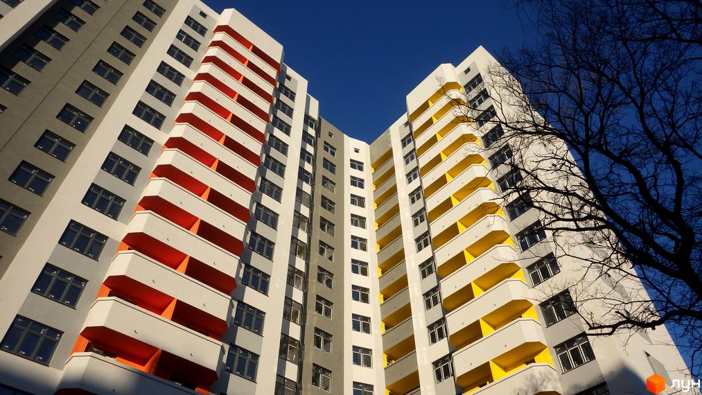 Багатоповерховий житловий комплекс Наш будинок з біло-сірим фасадом та яскравими помаранчевими і жовтими балконами на тлі синього неба.