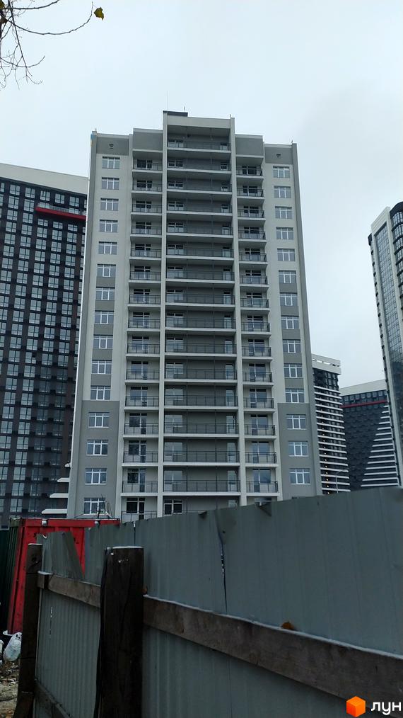 Фасад новобудови на вул. Причальна, 5а у Києві: висока будівля сірих та білих тонів з численними балконами та вікнами, за будівельним парканом.