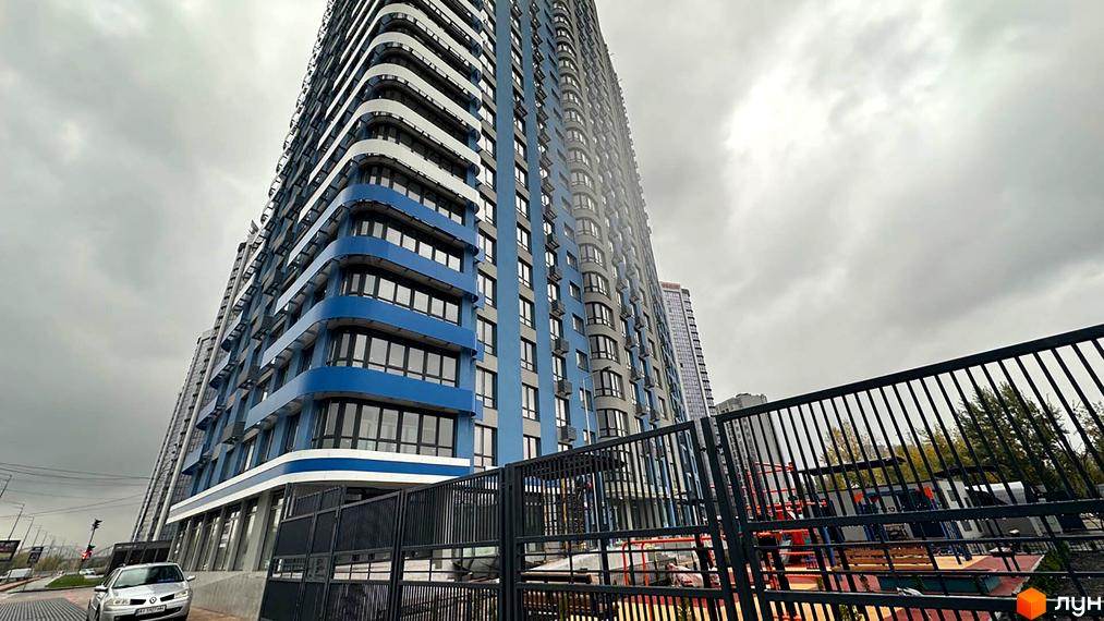 Зображення фасаду та прибудинкової території Житлового комплексу Берег Дніпра. Висотна будівля з синьо-білим оздобленням на фоні похмурого неба.