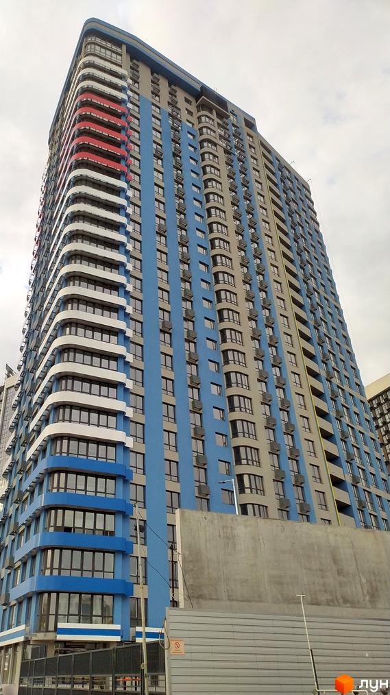 Зовнішній вигляд новобудови ЖК Берег Дніпра з блакитними та сірими фасадами, вигнутими балконами та великими вікнами. Навколо будинку видно будівельні роботи та паркан.