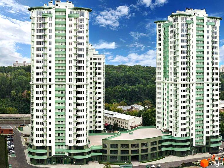 Житловий комплекс Смарагдовий: дві високі білі будівлі з зеленими елементами та численними вікнами. На передньому плані видно спільну основу, а на задньому – ліс та інші споруди.